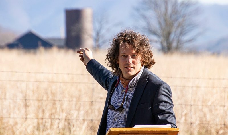 Adam Taylor points across a field of tall grass at the Catawba Sustainability Center while speaking at a podium outside.
