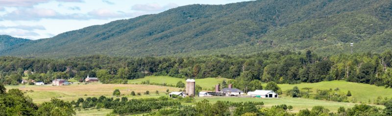 Green fields in the foreground with white farm buildings and two silos beyond surrounded by moutains.