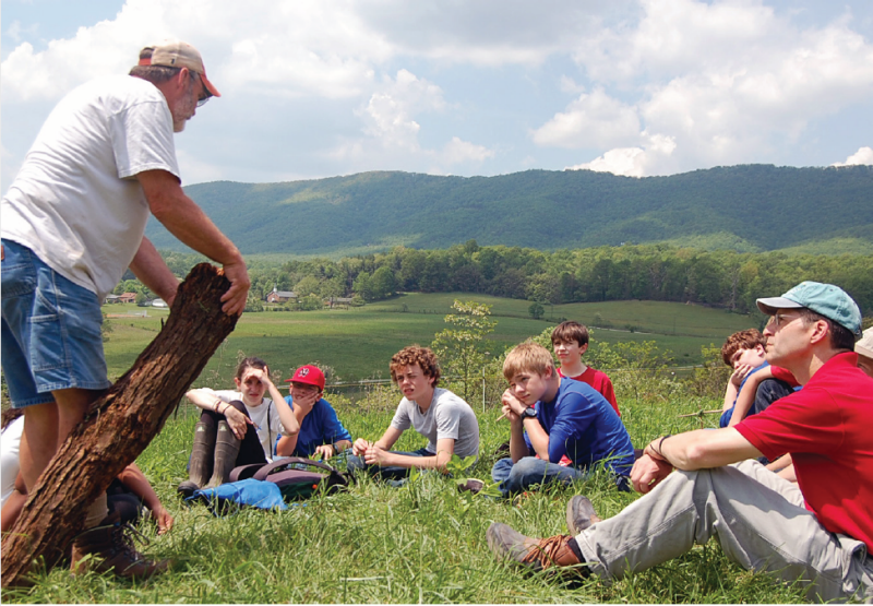 A man teaches to a group of children sitting in the grass high on a hill.