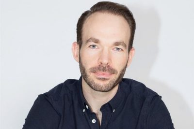 A man with short dark hair and a short beard, wearing a dark blue button-up shirt, looks into the camera, and smiles for a headshot.